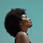 From below side view of young reflective ethnic female model with big earrings and closed eyes under blue sky